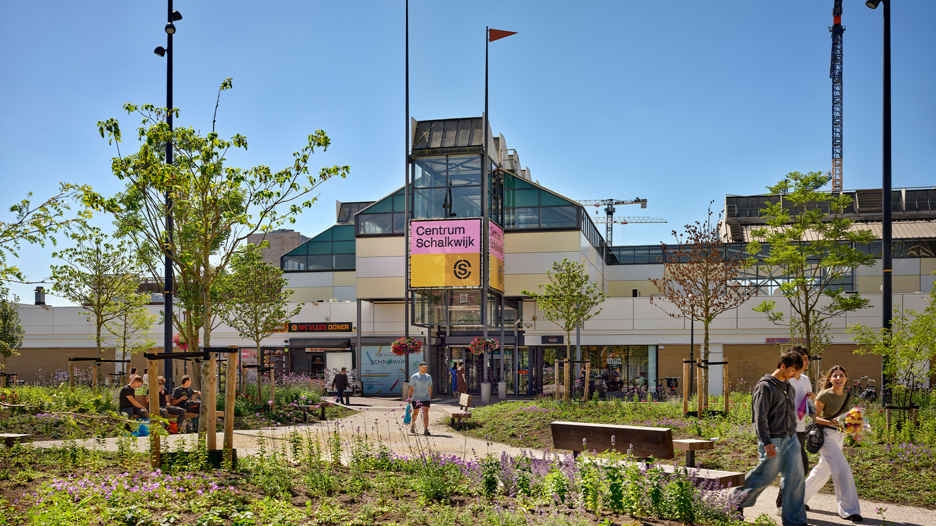 De ingang van een winkelcentrum waar verschillende mensen langslopen. Omringd met groen